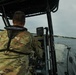 A military or law enforcement personnel wearing MultiCam/OCP camouflage uniform and a First Watch life vest is operating a rigid inflatable boat (RIB) on open water under overcast skies. The operator is seated at the helm console, which is equipped with a Raymarine navigation/chartplotter display and other electronic equipment. A distant shoreline with structures is visible on the horizon.