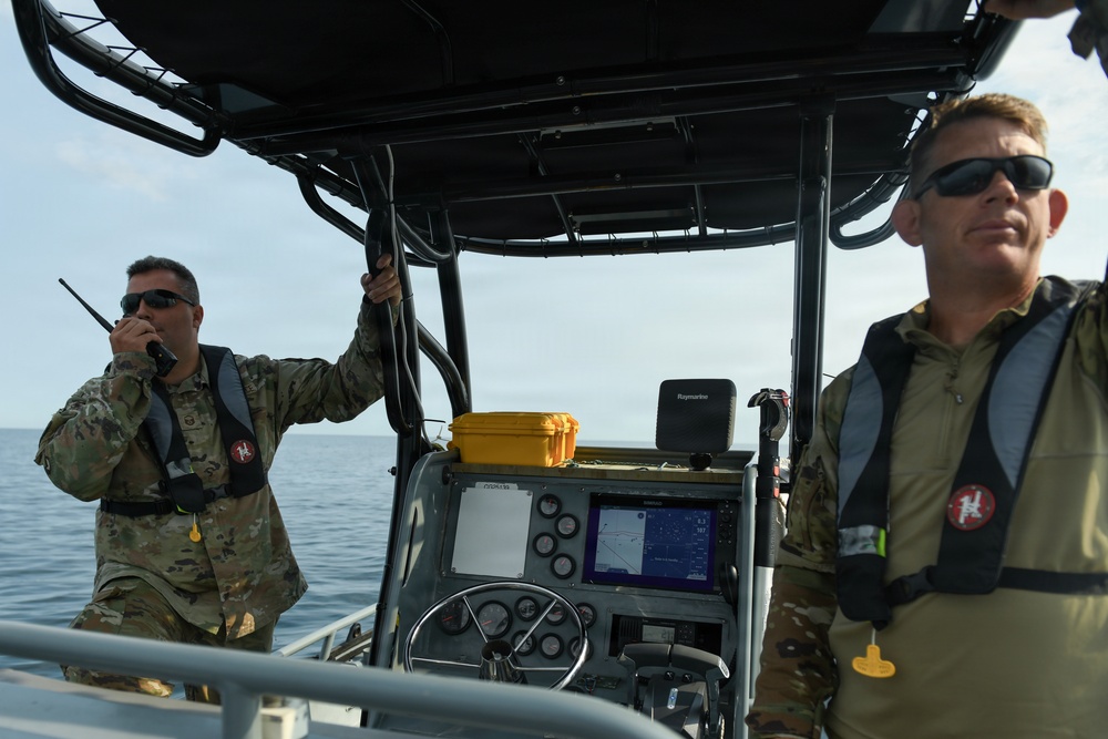 Two military personnel are aboard a patrol or utility boat on open water. One individual is communicating via handheld radio while gripping a support bar, and the other appears to be monitoring the surroundings. Both are wearing camouflage uniforms, inflatable life vests, and sunglasses.