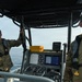 Two military personnel are aboard a patrol or utility boat on open water. One individual is communicating via handheld radio while gripping a support bar, and the other appears to be monitoring the surroundings. Both are wearing camouflage uniforms, inflatable life vests, and sunglasses.