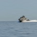 A military or law enforcement patrol vessel is traveling at high speed across calm, open water under a clear, hazy sky. The boat is producing a significant white wake and spray behind it, indicating considerable velocity. Several personnel are visible on the deck and bridge of the vessel.
