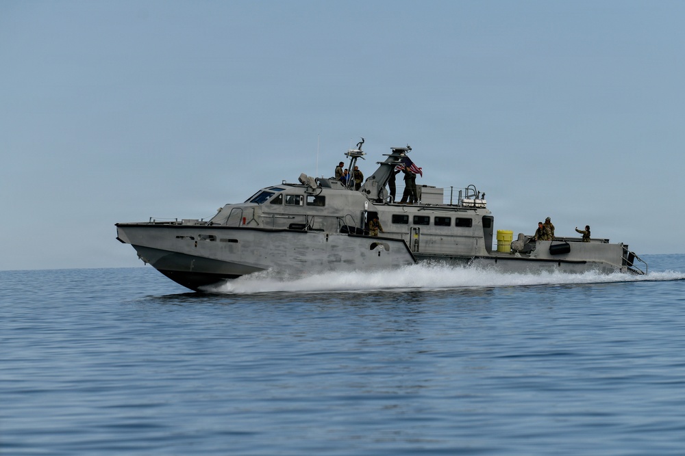 A gray military patrol boat is underway at speed on open water, generating a white wake along its hull. The vessel is flying an American flag and has several personnel visible on deck in military-style attire. The boat appears to be a U.S. Navy or special operations craft conducting an operational or training mission.