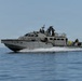A gray military patrol boat is underway at speed on open water, generating a white wake along its hull. The vessel is flying an American flag and has several personnel visible on deck in military-style attire. The boat appears to be a U.S. Navy or special operations craft conducting an operational or training mission.