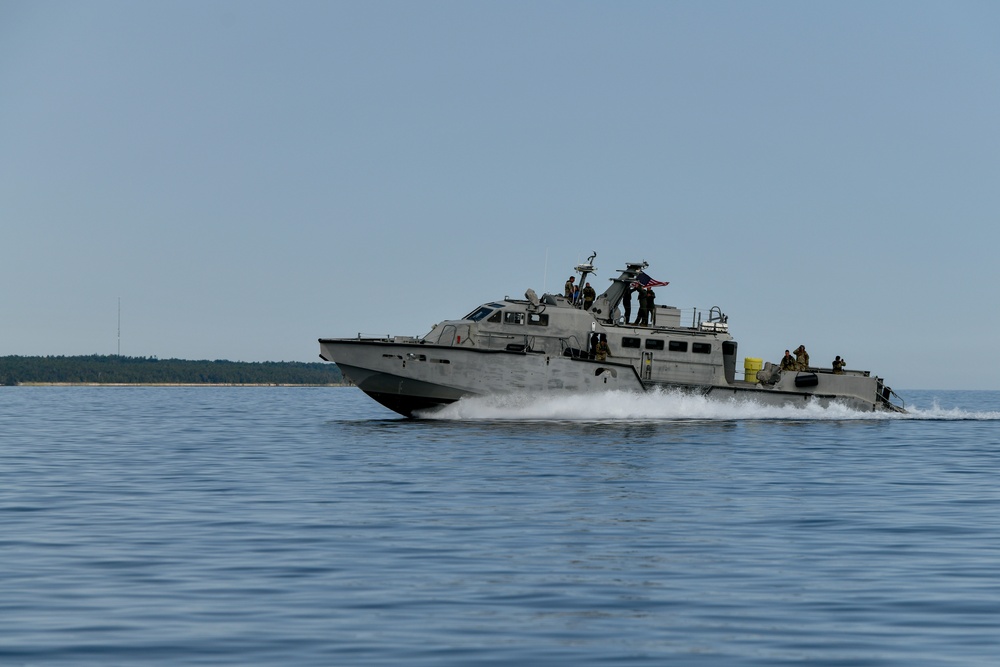 A gray military patrol boat is traveling at high speed across calm open water, generating a visible white wake. Several personnel in military attire are visible on deck, and a flag is flying from the vessel. A forested coastline is visible in the background under clear skies.