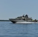 A gray military patrol boat is traveling at high speed across calm open water, generating a visible white wake. Several personnel in military attire are visible on deck, and a flag is flying from the vessel. A forested coastline is visible in the background under clear skies.