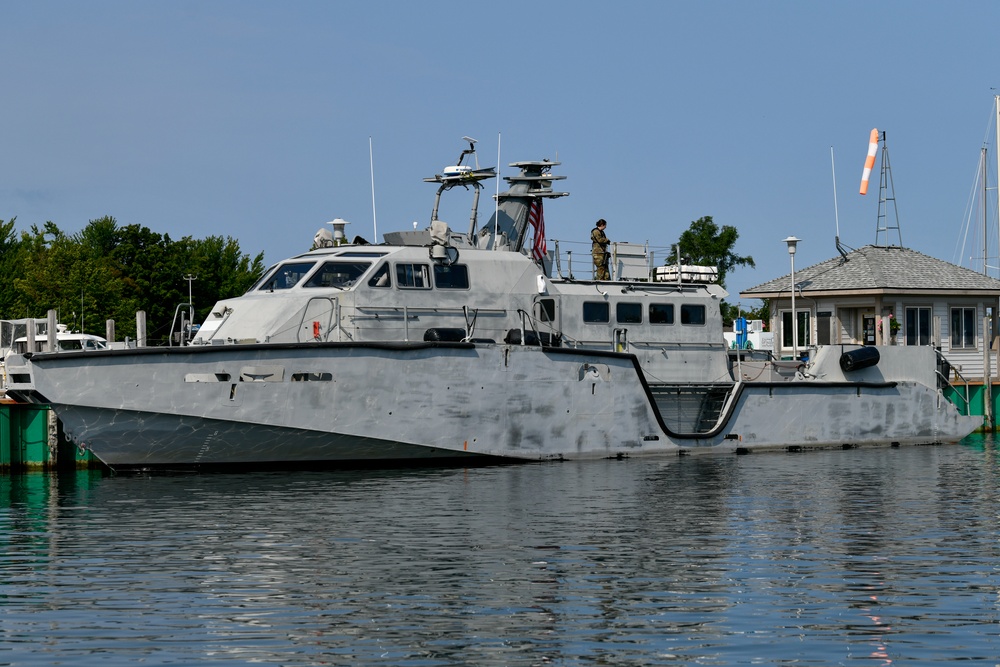 A gray military patrol vessel is docked at a marina in calm water conditions. At least one uniformed personnel is visible standing on the upper deck near the stern. The vessel features a modern angular hull design with various sensor and communication equipment mounted on the superstructure.