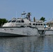 A gray military patrol vessel is docked at a marina in calm water conditions. At least one uniformed personnel is visible standing on the upper deck near the stern. The vessel features a modern angular hull design with various sensor and communication equipment mounted on the superstructure.