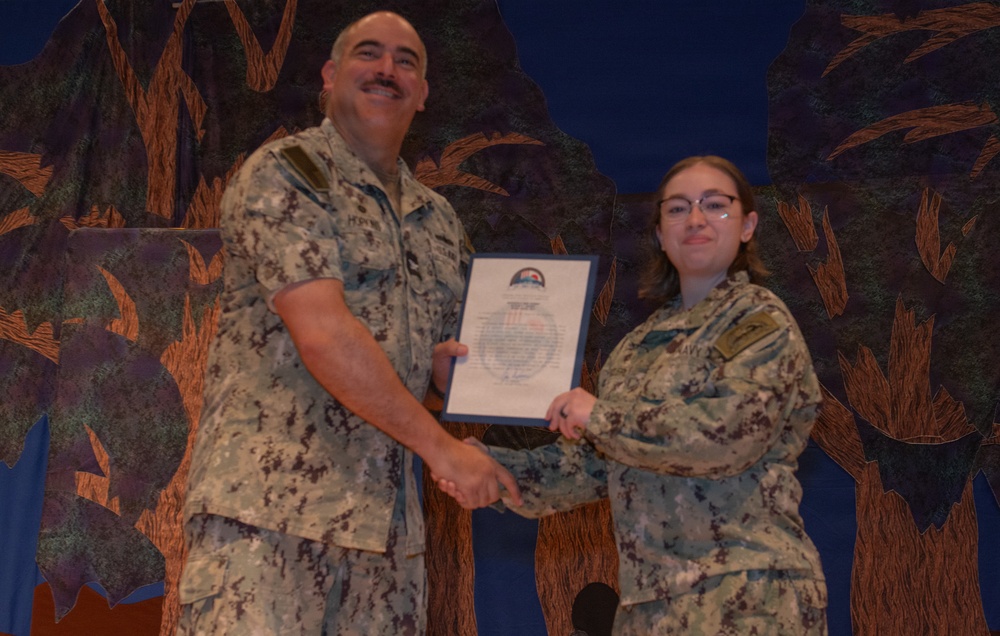Two individuals in military camouflage uniforms are posing together while jointly holding a framed certificate or award document. The ceremony appears to be a formal military recognition event, with the senior personnel presenting the award to the junior member. Both individuals are smiling, suggesting a positive occasion such as a commendation or achievement recognition.