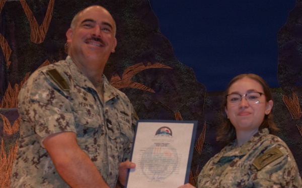 Two individuals in military camouflage uniforms are posing together while jointly holding a framed certificate or award document. The ceremony appears to be a formal military recognition event, with the senior personnel presenting the award to the junior member. Both individuals are smiling, suggesting a positive occasion such as a commendation or achievement recognition.