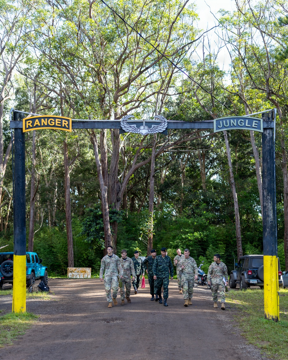 A group of military personnel in various camouflage uniforms walk through an entrance gate marked 'RANGER' on the left and 'JUNGLE' on the right, with an Airborne wings insignia centered at the top. The setting appears to be a military training facility surrounded by dense tropical or subtropical trees. The group includes personnel in what appear to be both U.S. Army and foreign military uniforms, suggesting a multinational training event.