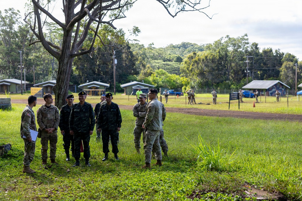 A group of military personnel from at least two different nations are gathered on a grassy field, appearing to be engaged in a briefing or discussion. Some soldiers wear US Army multicam uniforms while others wear darker military uniforms consistent with a Southeast Asian military force. The setting appears to be a military training area or base with small structures and additional personnel visible in the background.