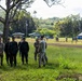 A group of military personnel from at least two different nations are gathered on a grassy field, appearing to be engaged in a briefing or discussion. Some soldiers wear US Army multicam uniforms while others wear darker military uniforms consistent with a Southeast Asian military force. The setting appears to be a military training area or base with small structures and additional personnel visible in the background.