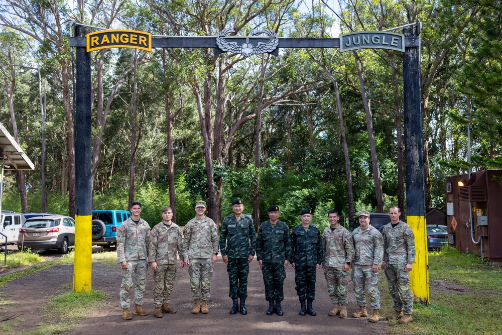 A group of nine military personnel pose for a formal group photograph under an archway gate marked 'RANGER' on the left and 'JUNGLE' on the right, with a military airborne/parachutist badge emblem at the top center. The group appears to include both U.S. Army soldiers in MultiCam/OCP uniforms and foreign military personnel in darker camouflage uniforms. The setting appears to be a military training facility entrance, likely the Jungle Operations Training Center in Hawaii.