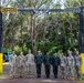 A group of nine military personnel pose for a formal group photograph under an archway gate marked 'RANGER' on the left and 'JUNGLE' on the right, with a military airborne/parachutist badge emblem at the top center. The group appears to include both U.S. Army soldiers in MultiCam/OCP uniforms and foreign military personnel in darker camouflage uniforms. The setting appears to be a military training facility entrance, likely the Jungle Operations Training Center in Hawaii.