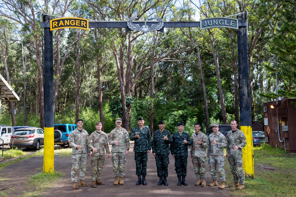 A group of nine military personnel pose for a group photo beneath a black arch gate displaying 'RANGER' and 'JUNGLE' tabs with an airborne wings emblem at the center. The group includes both U.S. Army soldiers in MultiCam/OCP uniforms and what appear to be Thai military officers in distinctive dark green camouflage uniforms. The setting appears to be a jungle warfare or ranger training facility, likely in Hawaii based on the vegetation.