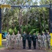 A group of nine military personnel pose for a group photo beneath a black arch gate displaying 'RANGER' and 'JUNGLE' tabs with an airborne wings emblem at the center. The group includes both U.S. Army soldiers in MultiCam/OCP uniforms and what appear to be Thai military officers in distinctive dark green camouflage uniforms. The setting appears to be a jungle warfare or ranger training facility, likely in Hawaii based on the vegetation.