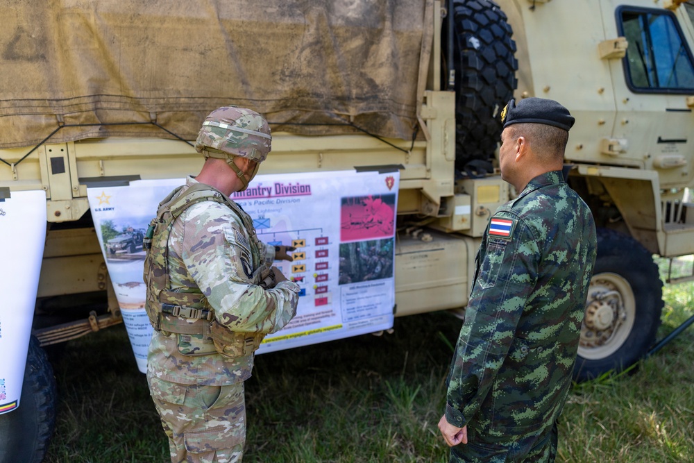 Two military personnel — one in U.S. Army multicam uniform and one in Thai military camouflage uniform with a Thai flag patch — are reviewing a briefing board mounted on a military vehicle. The board references the U.S. Army Infantry Division and appears to be part of a joint military exercise or liaison event. The setting is an outdoor area with military vehicles present.