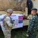 Two military personnel — one in U.S. Army multicam uniform and one in Thai military camouflage uniform with a Thai flag patch — are reviewing a briefing board mounted on a military vehicle. The board references the U.S. Army Infantry Division and appears to be part of a joint military exercise or liaison event. The setting is an outdoor area with military vehicles present.