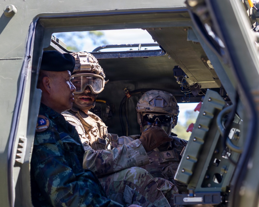 Three military personnel are seated inside what appears to be a military armored vehicle or tactical transport. Two soldiers wearing combat helmets with goggles and multicam/tan uniforms are engaged with a third individual wearing a blue camouflage uniform and black beret, suggesting a multinational or joint military interaction. The scene appears to involve a demonstration or familiarization of vehicle equipment.