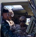 Three military personnel are seated inside what appears to be a military armored vehicle or tactical transport. Two soldiers wearing combat helmets with goggles and multicam/tan uniforms are engaged with a third individual wearing a blue camouflage uniform and black beret, suggesting a multinational or joint military interaction. The scene appears to involve a demonstration or familiarization of vehicle equipment.