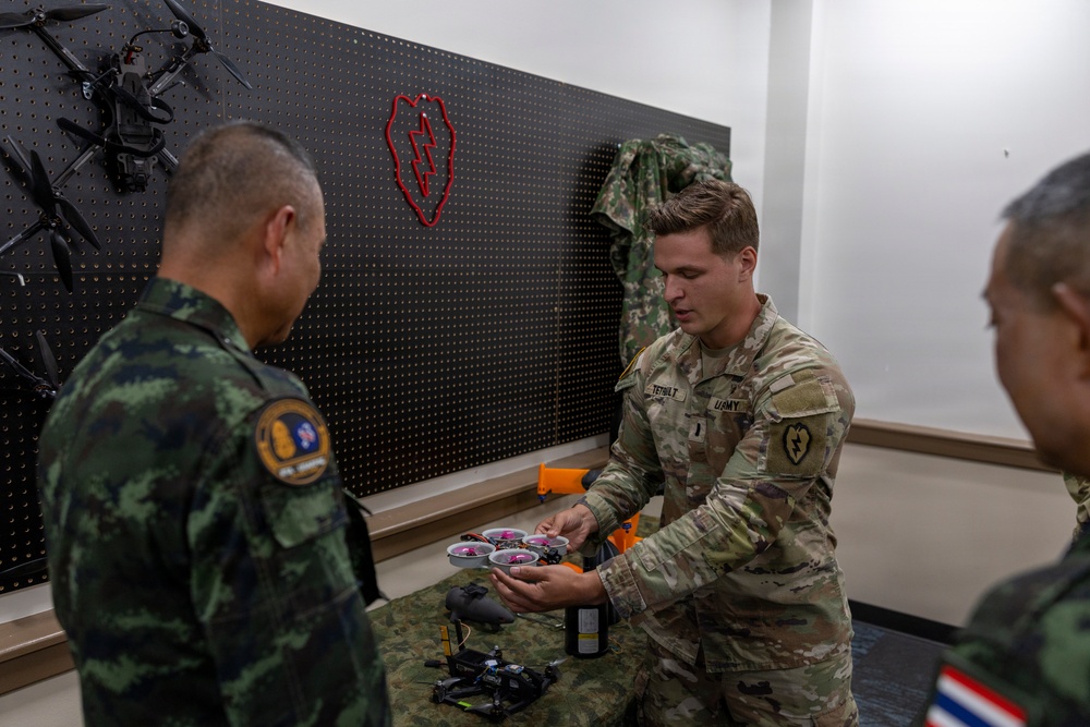 A U.S. Army soldier, identified by name tape as 'TERHILT' and wearing a 25th Infantry Division patch, demonstrates a small racing/FPV drone to what appear to be Thai military personnel in a drone equipment room. The setting appears to be an indoor briefing or demonstration area with multiple drones mounted on a pegboard wall. This appears to be a military-to-military exchange or joint training event.