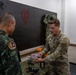 A U.S. Army soldier, identified by name tape as 'TERHILT' and wearing a 25th Infantry Division patch, demonstrates a small racing/FPV drone to what appear to be Thai military personnel in a drone equipment room. The setting appears to be an indoor briefing or demonstration area with multiple drones mounted on a pegboard wall. This appears to be a military-to-military exchange or joint training event.