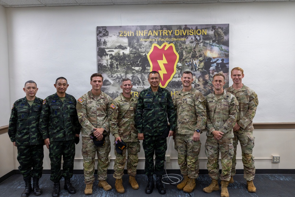 A group of eight military personnel pose for a formal group photo in front of a 25th Infantry Division banner reading 'America's Pacific Division.' The group includes both U.S. Army soldiers in OCP (Operational Camouflage Pattern) uniforms and foreign military personnel in distinctive camouflage uniforms, suggesting a multinational military engagement or visit. The setting appears to be an indoor briefing or conference room at a U.S. Army installation.