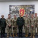A group of eight military personnel pose for a formal group photo in front of a 25th Infantry Division banner reading 'America's Pacific Division.' The group includes both U.S. Army soldiers in OCP (Operational Camouflage Pattern) uniforms and foreign military personnel in distinctive camouflage uniforms, suggesting a multinational military engagement or visit. The setting appears to be an indoor briefing or conference room at a U.S. Army installation.