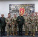 A group of eight military personnel pose for a formal group photo in front of a 25th Infantry Division banner reading 'America's Pacific Division.' The group includes both U.S. Army soldiers in OCP (Operational Camouflage Pattern) uniforms and foreign military personnel in different camouflage uniforms, suggesting a multinational military engagement or exchange. Several individuals are making the 'shaka' hand gesture, consistent with Hawaiian military culture associated with the 25th Infantry Division based at Schofield Barracks, Hawaii.