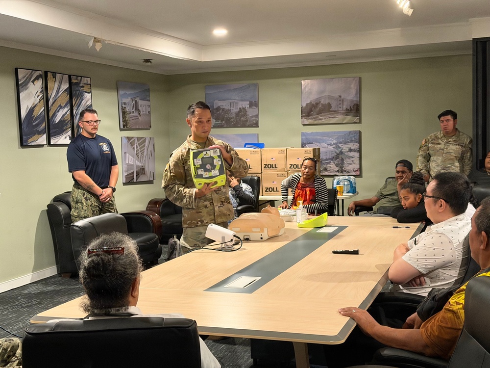 A military service member in camouflage uniform is conducting a training session, holding up a ZOLL AED (Automated External Defibrillator) device for a group of seated attendees in a conference room. Multiple boxes labeled 'ZOLL' are visible in the background, suggesting a distribution or training event. A second person in a navy uniform stands nearby, and another soldier in camouflage stands at the back of the room.