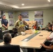 A military service member in camouflage uniform is conducting a training session, holding up a ZOLL AED (Automated External Defibrillator) device for a group of seated attendees in a conference room. Multiple boxes labeled 'ZOLL' are visible in the background, suggesting a distribution or training event. A second person in a navy uniform stands nearby, and another soldier in camouflage stands at the back of the room.