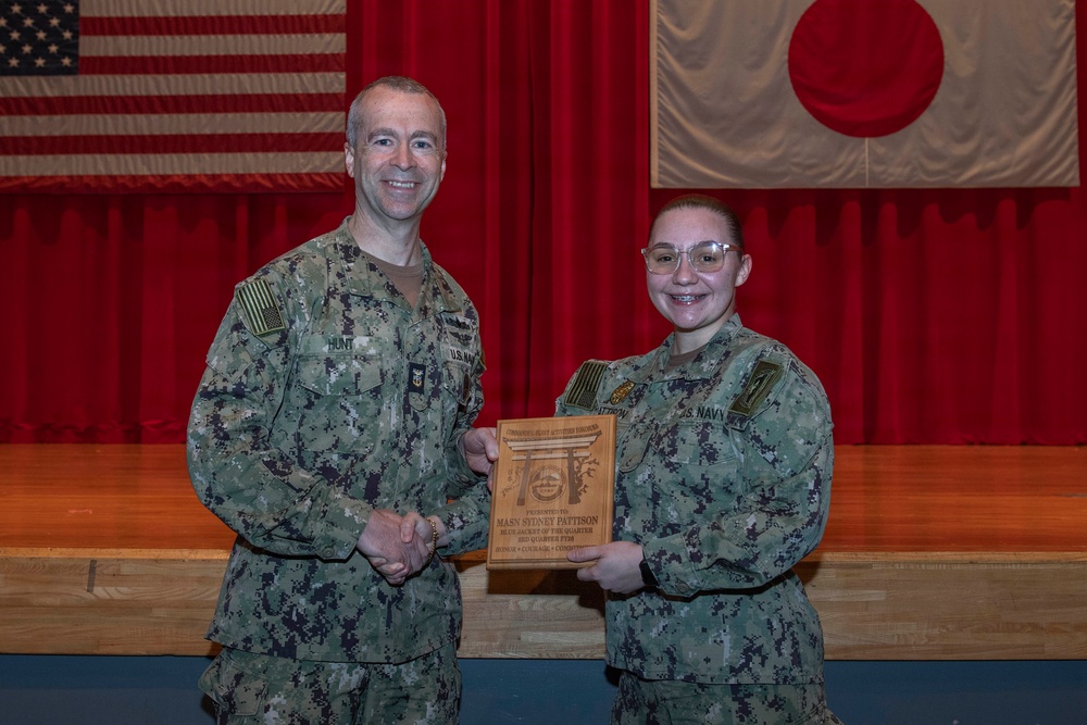 Two U.S. Navy personnel in NWU Type III (digital camouflage) uniforms pose together holding a wooden award plaque on a stage. The plaque appears to be a 'Bluejacket of the Quarter' award presented to MASN Sydney Pattison. The setting appears to be a military installation in Japan, evidenced by the American and Japanese flags displayed on the red-curtained stage backdrop.