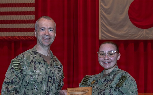 Two U.S. Navy personnel in NWU Type III (digital camouflage) uniforms pose together holding a wooden award plaque on a stage. The plaque appears to be a 'Bluejacket of the Quarter' award presented to MASN Sydney Pattison. The setting appears to be a military installation in Japan, evidenced by the American and Japanese flags displayed on the red-curtained stage backdrop.