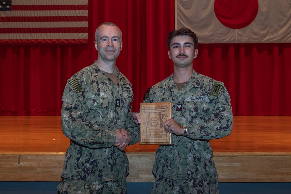 Two U.S. Navy personnel in NWU Type III (AOR-2) camouflage uniforms are posing together on a stage during what appears to be an awards ceremony. The sailor on the right is holding a wooden plaque/award while both individuals display it for the photo. An American flag and Japanese flag are visible in the background, suggesting this ceremony is taking place at a U.S. military installation in Japan.