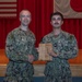 Two U.S. Navy personnel in NWU Type III (AOR-2) camouflage uniforms are posing together on a stage during what appears to be an awards ceremony. The sailor on the right is holding a wooden plaque/award while both individuals display it for the photo. An American flag and Japanese flag are visible in the background, suggesting this ceremony is taking place at a U.S. military installation in Japan.