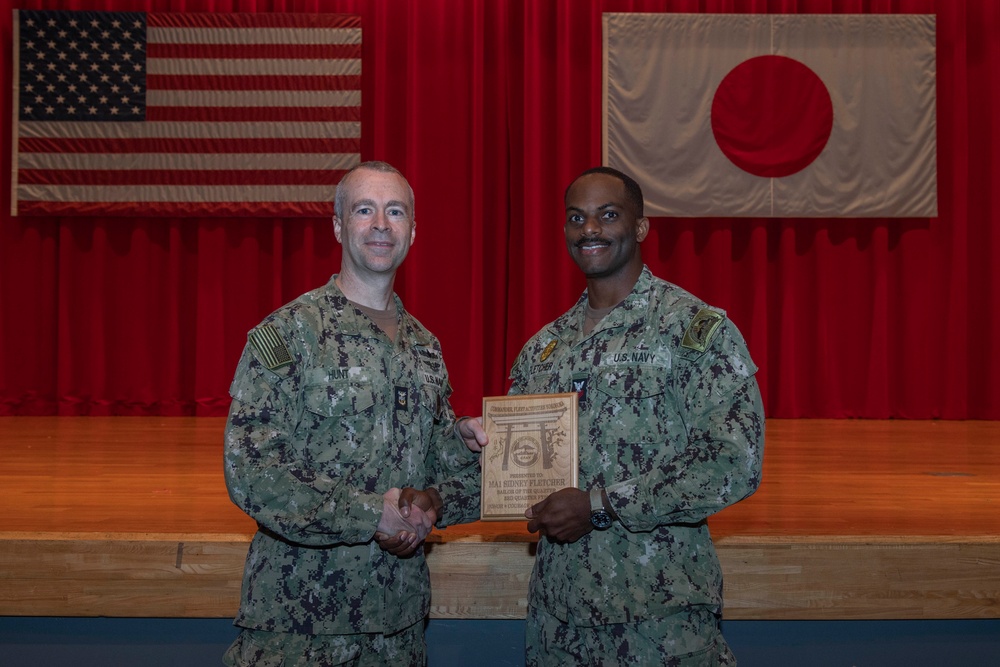 Two U.S. Navy personnel in NWU Type III (digital camouflage) uniforms pose together holding a wooden award plaque. The ceremony appears to take place in a auditorium or gymnasium setting with an American flag and Japanese flag displayed on the stage backdrop. Based on visible name tapes and the plaque inscription, this appears to be an award presentation to MA1 Sidney Fletcher.