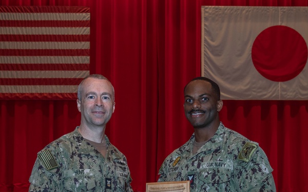 Two U.S. Navy personnel in NWU Type III (digital camouflage) uniforms pose together holding a wooden award plaque. The ceremony appears to take place in a auditorium or gymnasium setting with an American flag and Japanese flag displayed on the stage backdrop. Based on visible name tapes and the plaque inscription, this appears to be an award presentation to MA1 Sidney Fletcher.