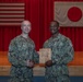 Two U.S. Navy personnel in NWU Type III (digital camouflage) uniforms pose together holding a wooden award plaque. The ceremony appears to take place in a auditorium or gymnasium setting with an American flag and Japanese flag displayed on the stage backdrop. Based on visible name tapes and the plaque inscription, this appears to be an award presentation to MA1 Sidney Fletcher.