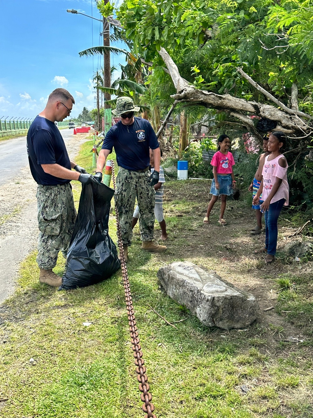 Two U.S. Navy personnel in digital camouflage uniforms are conducting a community cleanup operation along a roadside, collecting debris into a large black trash bag. Several local children, appearing to be girls, watch the cleanup activity with interest. The setting appears to be a tropical location based on the vegetation and environmental conditions.