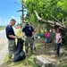 Two U.S. Navy personnel in digital camouflage uniforms are conducting a community cleanup operation along a roadside, collecting debris into a large black trash bag. Several local children, appearing to be girls, watch the cleanup activity with interest. The setting appears to be a tropical location based on the vegetation and environmental conditions.
