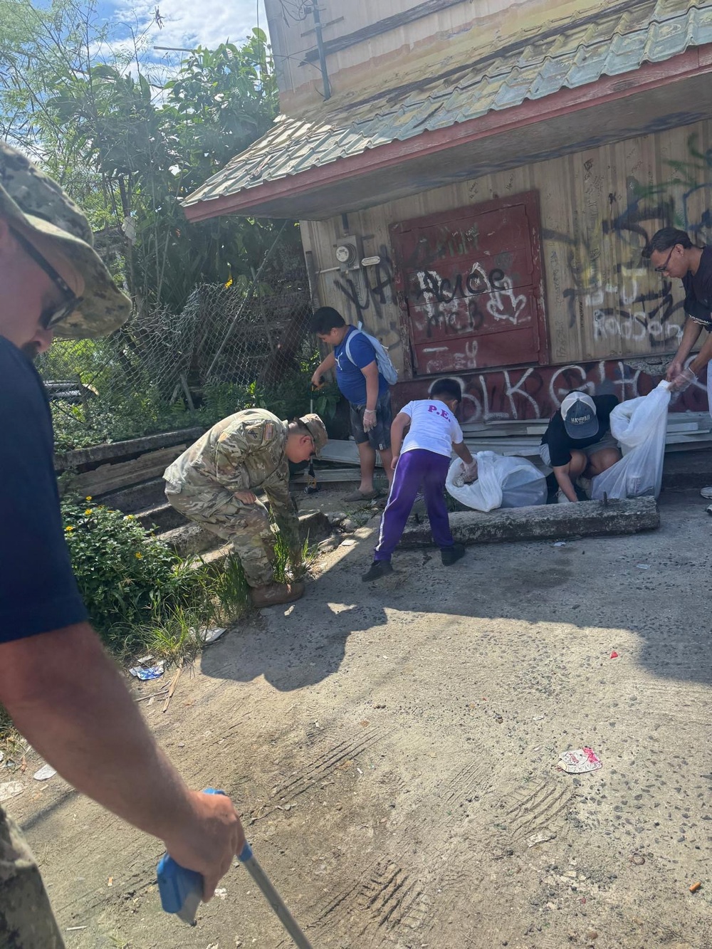 A group of individuals, including at least one person in military camouflage uniform, are engaged in a community cleanup effort near a dilapidated building covered in graffiti. Participants are collecting debris and trash into large white garbage bags in what appears to be a neglected urban area. The scene suggests an organized volunteer or civic service activity.