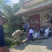 A group of individuals, including at least one person in military camouflage uniform, are engaged in a community cleanup effort near a dilapidated building covered in graffiti. Participants are collecting debris and trash into large white garbage bags in what appears to be a neglected urban area. The scene suggests an organized volunteer or civic service activity.