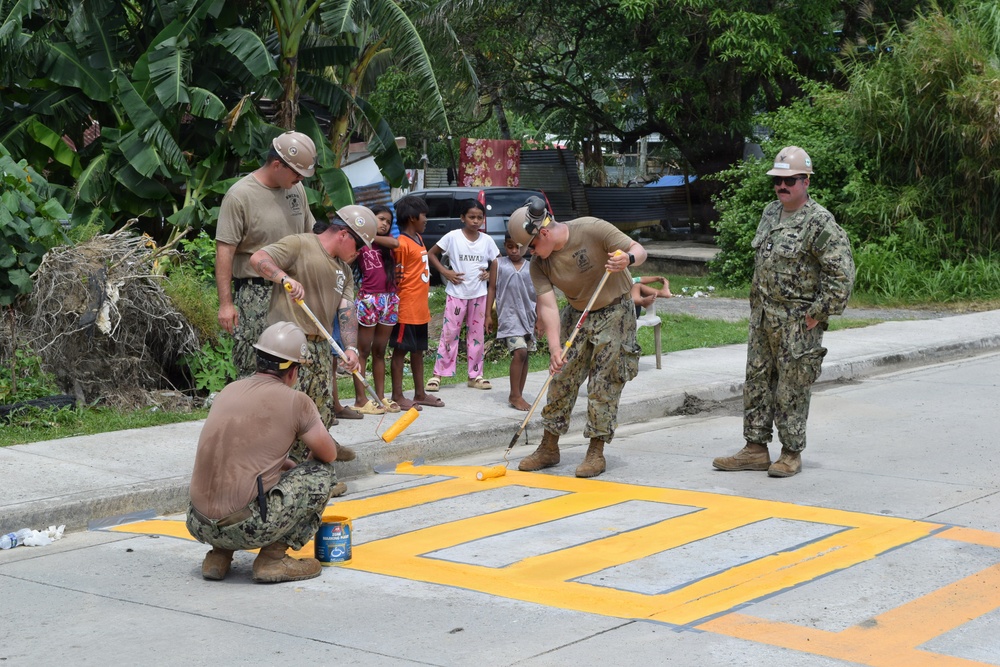 U.S. Navy personnel in uniform are painting yellow road markings, likely a pedestrian crosswalk, on a concrete street in what appears to be a tropical location. Several sailors wearing helmets and tan t-shirts with Navy camouflage trousers are using paint rollers, while a senior officer in full camouflage uniform observes. A group of local children watches the work from the sidewalk, suggesting this is a community assistance or humanitarian mission.