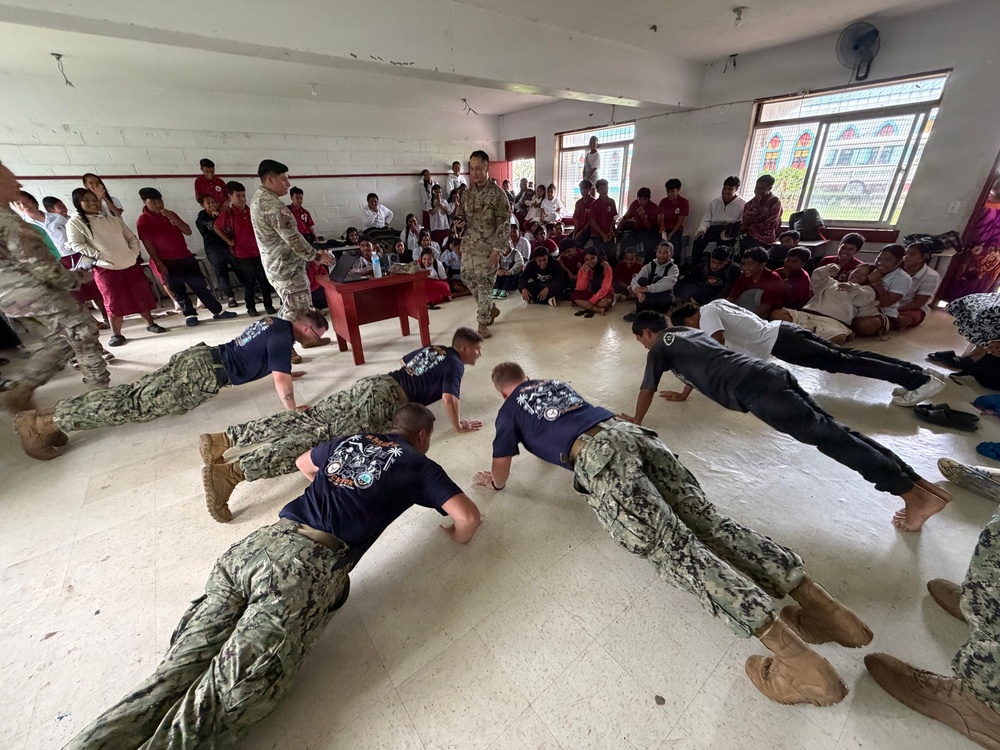 Military personnel in camouflage uniforms and navy blue t-shirts are performing push-ups on the floor of what appears to be a school or community hall, while two uniformed supervisors stand and observe. A large crowd of students and civilians, many wearing red and white clothing, watch from the sides and background. This appears to be a military community outreach or demonstration event at an educational institution.