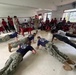 Military personnel in camouflage uniforms and navy blue t-shirts are performing push-ups on the floor of what appears to be a school or community hall, while two uniformed supervisors stand and observe. A large crowd of students and civilians, many wearing red and white clothing, watch from the sides and background. This appears to be a military community outreach or demonstration event at an educational institution.