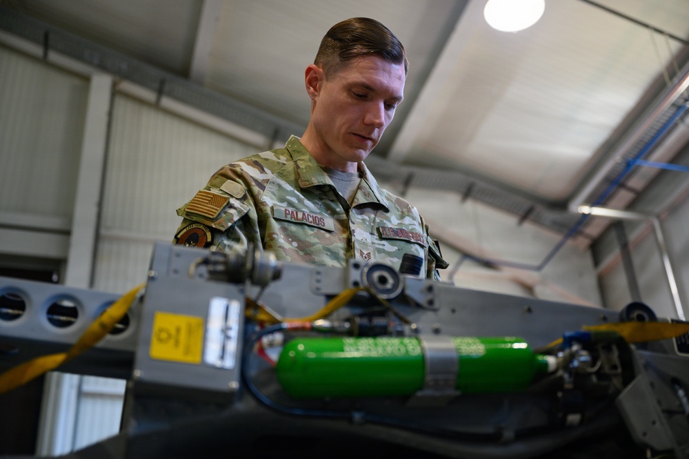 A U.S. Air Force service member named Palacios, wearing OCP (Operational Camouflage Pattern) uniform, is working on military equipment in what appears to be a maintenance hangar or workshop. The individual is focused on inspecting or servicing a piece of equipment that includes yellow cables, a green cylindrical component, and various mechanical parts. The setting suggests an aircraft or weapons system maintenance environment.