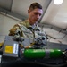 A U.S. Air Force service member named Palacios, wearing OCP (Operational Camouflage Pattern) uniform, is working on military equipment in what appears to be a maintenance hangar or workshop. The individual is focused on inspecting or servicing a piece of equipment that includes yellow cables, a green cylindrical component, and various mechanical parts. The setting suggests an aircraft or weapons system maintenance environment.