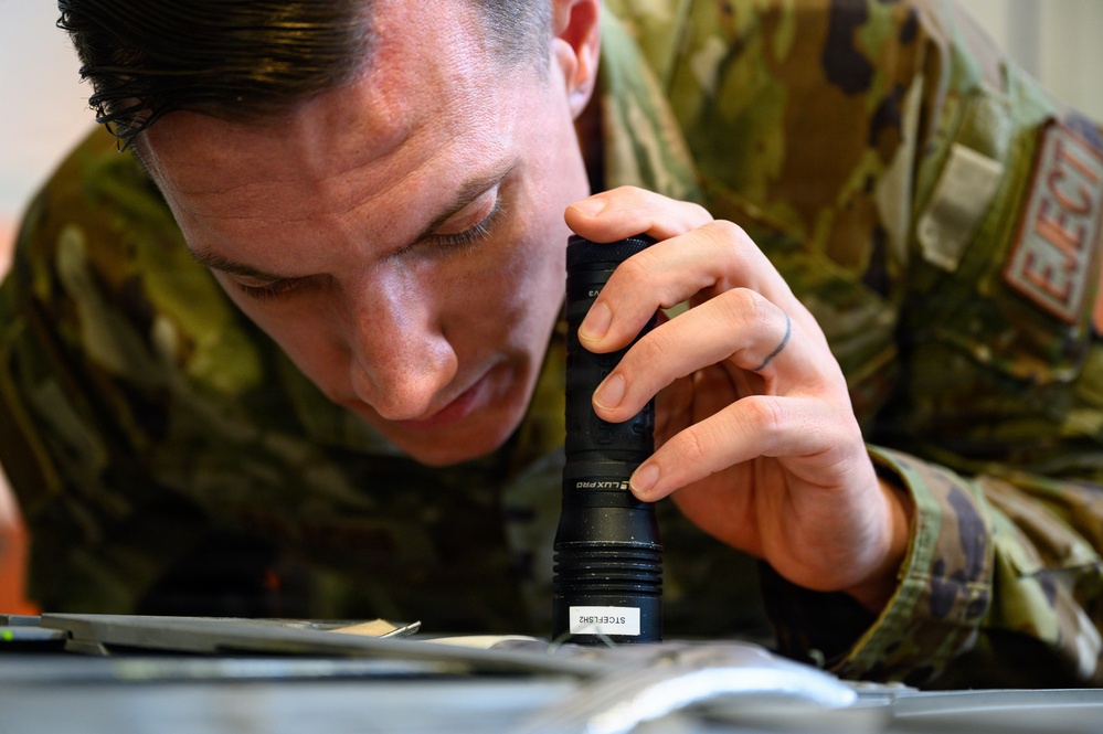 A military service member in OCP (Operational Camouflage Pattern) uniform is closely inspecting a surface or component using a LuxPro brand flashlight. The individual is leaning in very close to examine what appears to be a piece of equipment or documentation. A red patch with the word 'EJECT' is visible on the uniform sleeve, suggesting possible aircrew or aviation maintenance association.