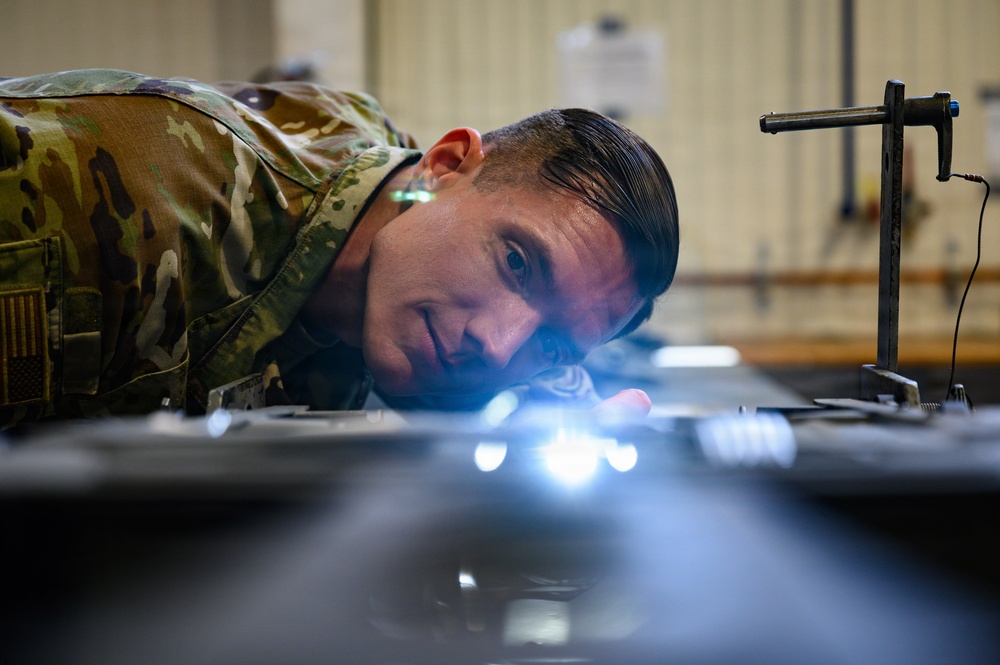 A military service member in OCP (Operational Camouflage Pattern) uniform is closely inspecting a surface or component at eye level, using a bright light source to examine fine details. The individual appears to be conducting a precision inspection or quality check in what appears to be a maintenance or technical facility. The low-angle perspective and intense focus suggest a detailed technical inspection task.