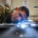 A military service member in OCP (Operational Camouflage Pattern) uniform is closely inspecting a surface or component at eye level, using a bright light source to examine fine details. The individual appears to be conducting a precision inspection or quality check in what appears to be a maintenance or technical facility. The low-angle perspective and intense focus suggest a detailed technical inspection task.