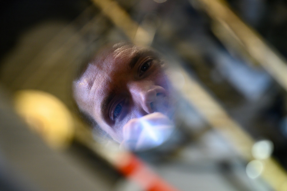 A close-up, low-angle photograph of a man's face reflected or viewed through a glass or transparent surface, illuminated by a bright light source. The image is taken from an unusual perspective, likely looking up through a glass container or optical element. The composition creates a distorted, artistic view of the subject examining or inspecting something closely.